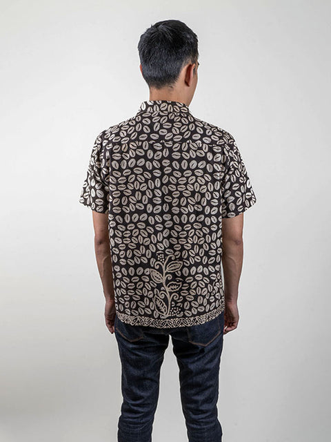 A person wearing a black shirt looking from the back made from traditional Indonesian batik fabric. The shirt is gender-neutral and has a loose fit. The focus is on the shirt and its intricate batik pattern, which features a mix of black, gray, and white colors