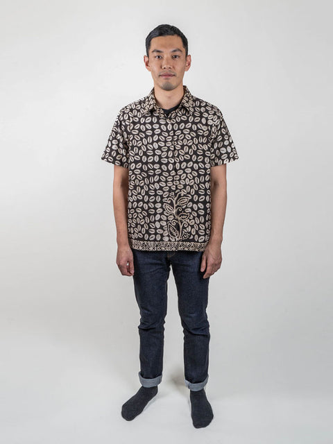 A person wearing a black shirt made from traditional Indonesian batik fabric. The shirt is gender-neutral and has a loose fit. The focus is on the shirt and its intricate batik pattern, which features a mix of black, gray, and white colors