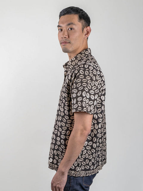 A person wearing a black shirt made from traditional Indonesian batik fabric. The shirt is gender-neutral and has a loose fit. The person is standing against a gray background and has a serious expression on their face. The focus is on the shirt and its intricate batik pattern